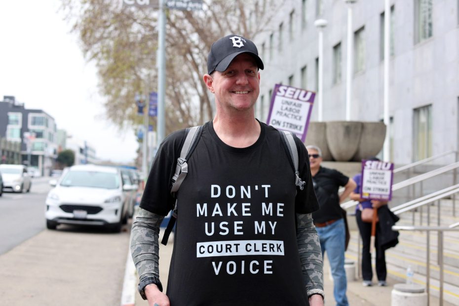 A man wearing a "Don't make me use my court clerk voice" T-shirt stands outside a building, smiling, with people holding "SEIU on strike" signs in the background.