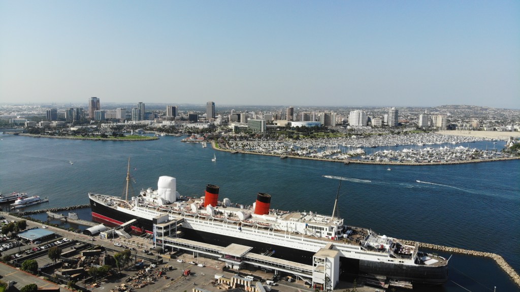 Aerial view of the Queen Mary ship docked at Long Beach harbor, with the city skyline and numerous boats in the marina in the background.