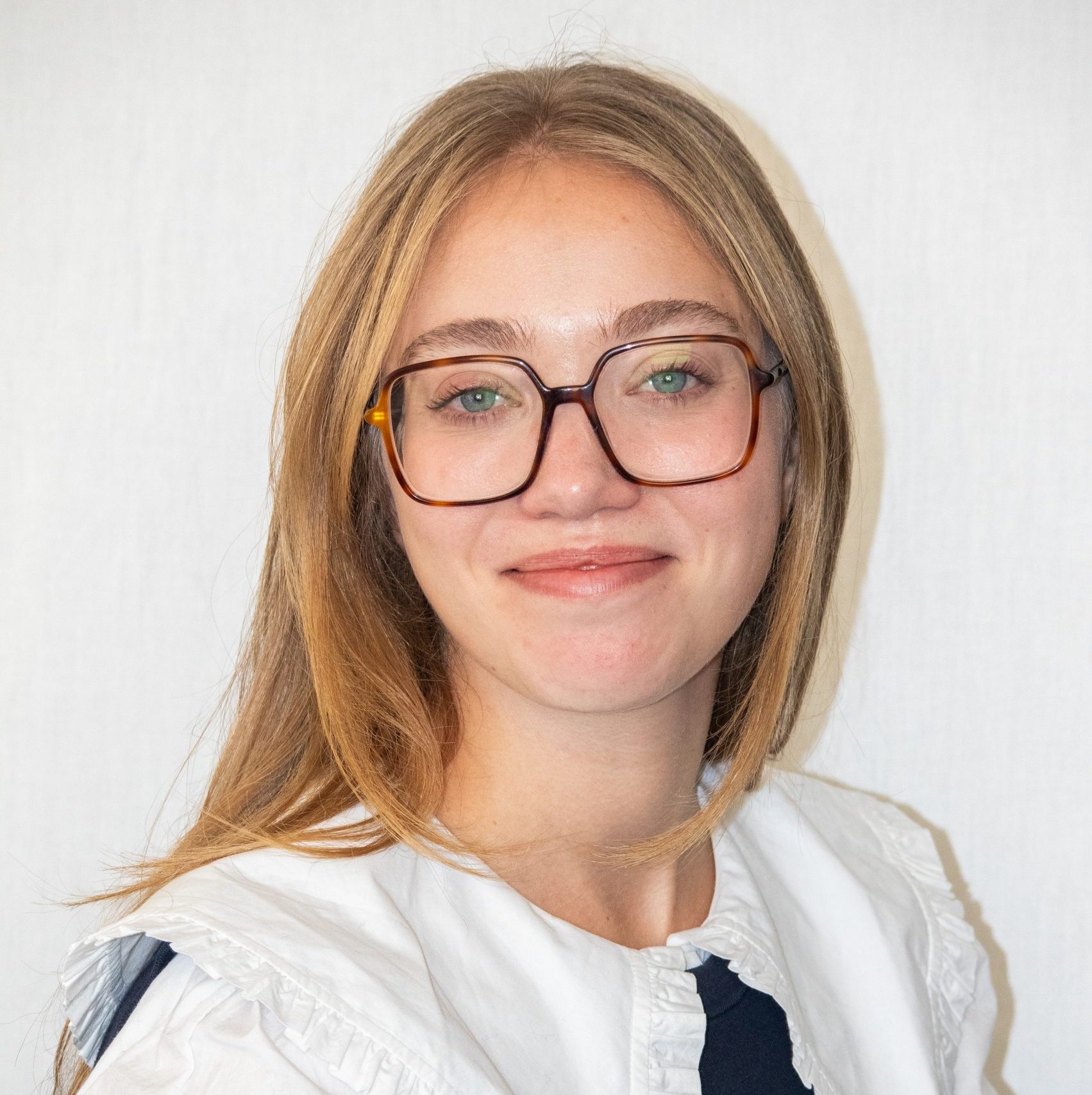 A woman with long blonde hair and large glasses, wearing a white ruffled top, smiles slightly in front of a plain light background.