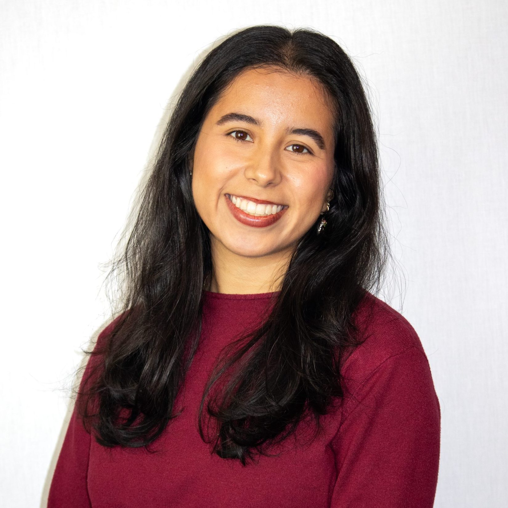 A woman with long dark hair wearing a maroon sweater stands smiling in front of a plain white background.