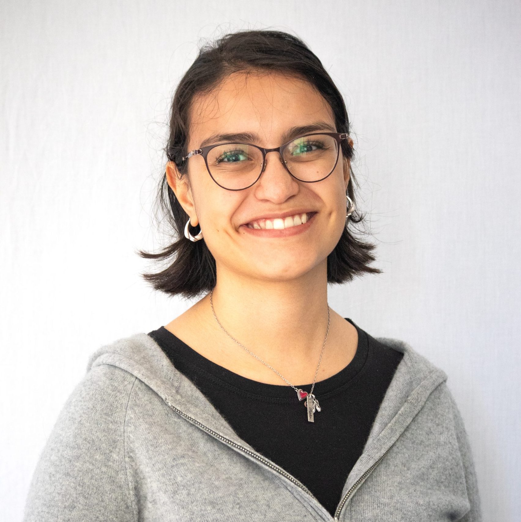 A person with short dark hair and glasses smiles at the camera. They are wearing a gray zip-up hoodie, black shirt, and a necklace. The background is plain and light-colored.