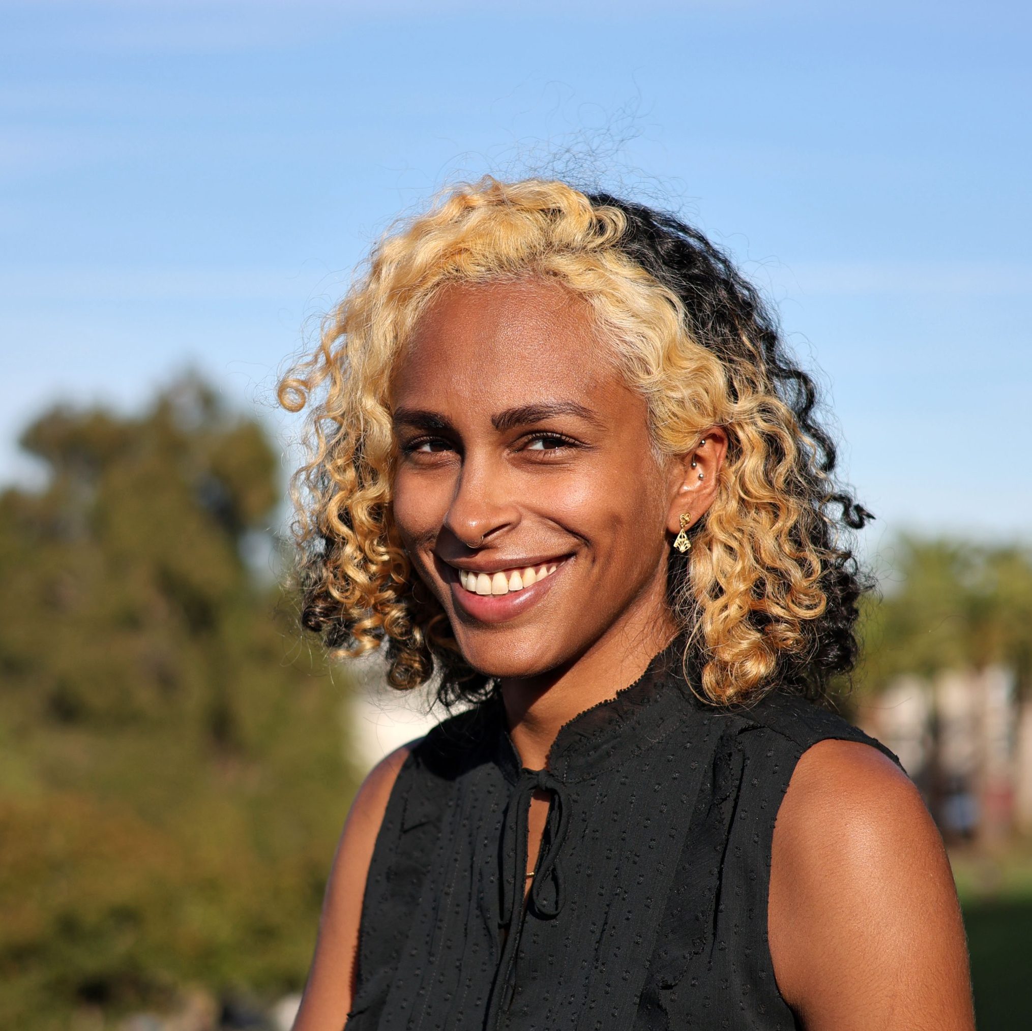 Smiling person with curly blonde and black hair, wearing a black sleeveless top, standing outdoors with trees and a clear sky in the background.