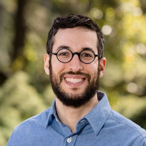 Man with beard and glasses poses outdoors in front of foliage