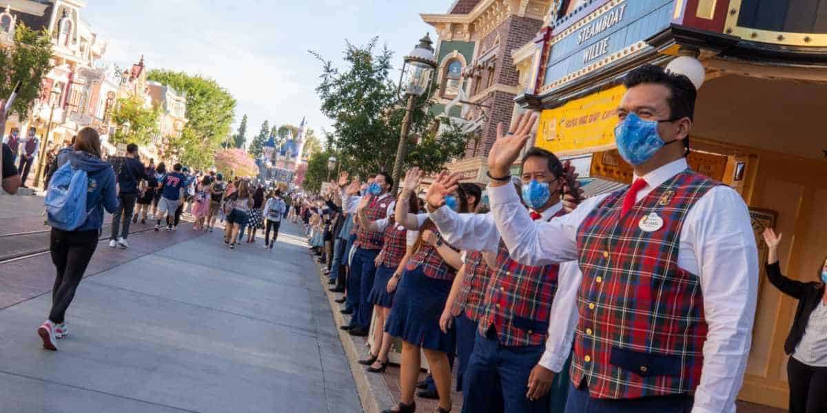 A group of Disneyland cast members, dressed in red and blue plaid vests and blue face masks, line both sides of a street, waving to guests who are walking towards the camera.