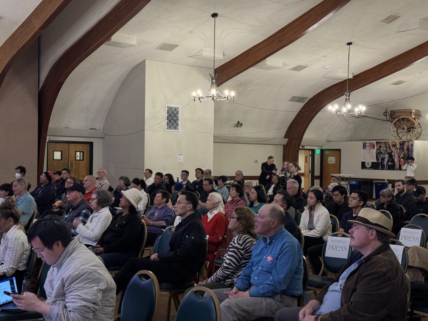 A group of people sit in rows of chairs in a hall with arched wooden beams, listening to a presentation or event.