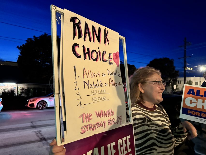 A person holds a sign explaining ranked choice voting, listing candidate options and stating "The winning strategy for DA" during an outdoor evening event.