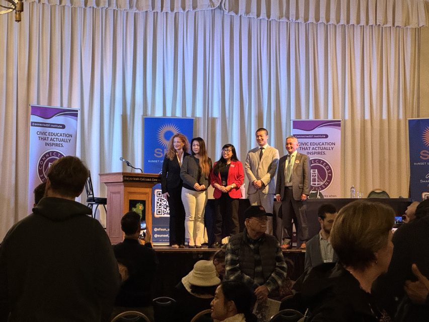 A group of five people pose on stage in front of banners that read "Civic Education That Factually Inspires" at an indoor event with an audience in the foreground.