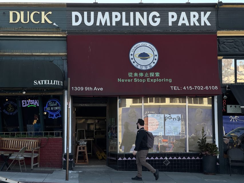 A person walks past Dumpling Park restaurant with a red awning displaying its name, logo, address, slogan, and phone number. Neighboring businesses are partially visible.