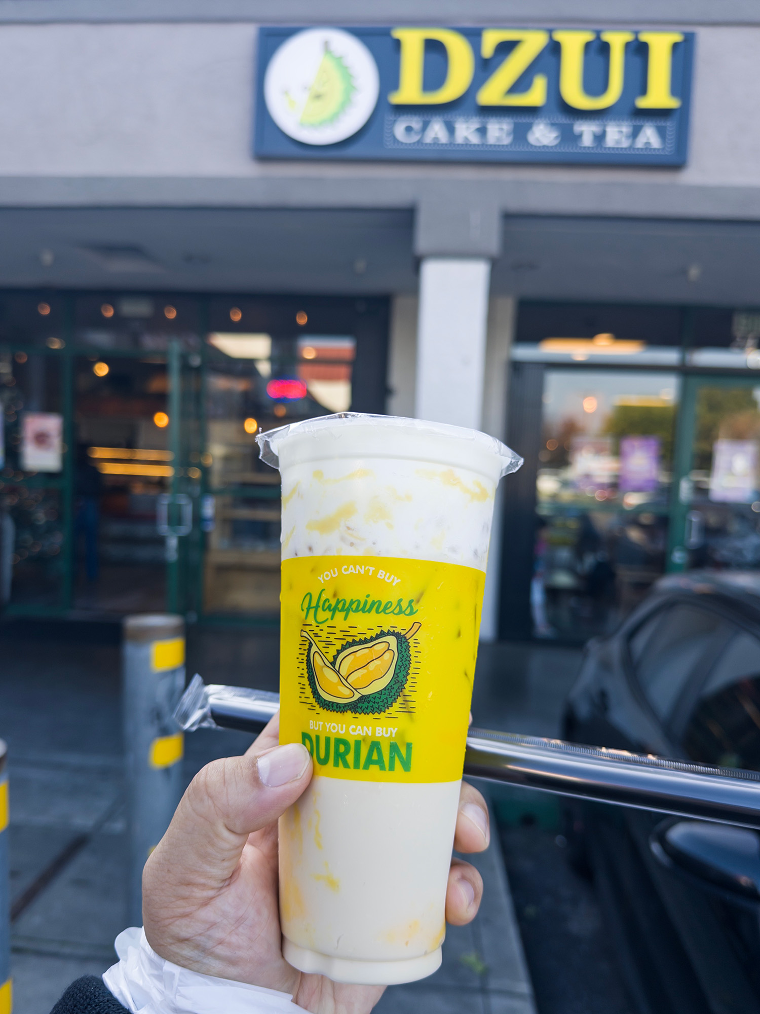 A hand holds a cup of milk tea. The label reads, "You can't buy happiness, but you can buy durian."