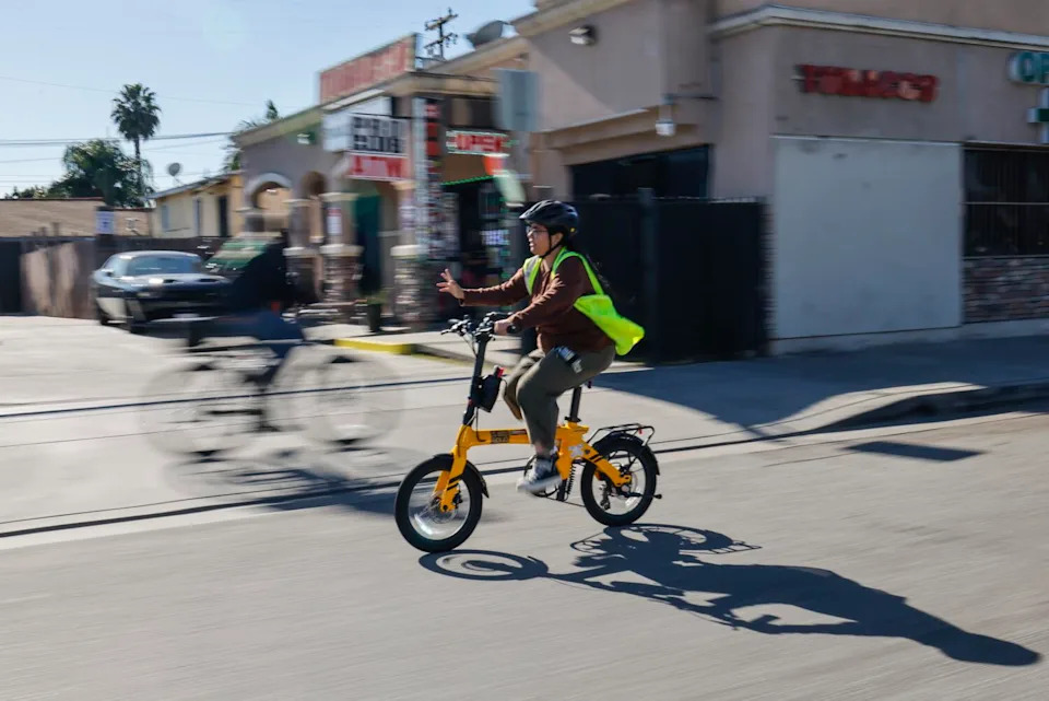 A woman waves while biking on a city street.