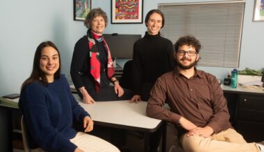 Four people smiling in an office setting, standing and sitting around a desk.