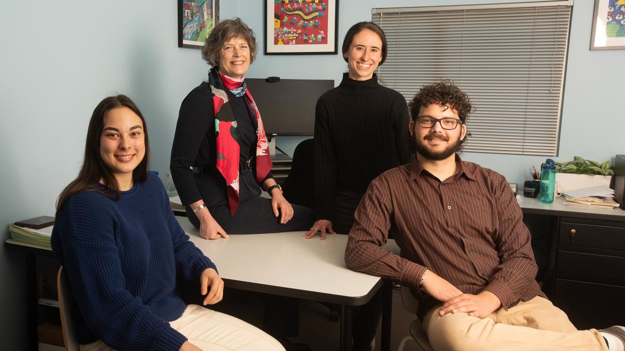 Four people smiling in an office setting, standing and sitting around a desk.