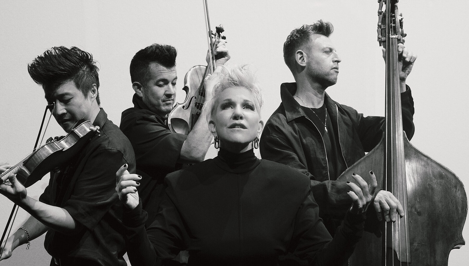 a dramatic black-and-white photo of Joyce DiDonato gently looking toward the sky with her hands lifting up, with the three strings musicians from the group Time for Three in the background.