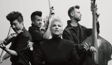 a dramatic black-and-white photo of Joyce DiDonato gently looking toward the sky with her hands lifting up, with the three strings musicians from the group Time for Three in the background.