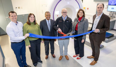 Five people in a room stand next to man with large scissors at ribbon-cutting ceremony with a cylindrical machine nearby.