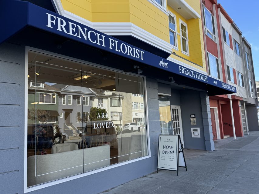 Street view of a yellow and gray building with a navy blue awning labeled "French Florist." A sign in the window reads "YOU ARE LOVED" and a sidewalk sign says "NOW OPEN!.