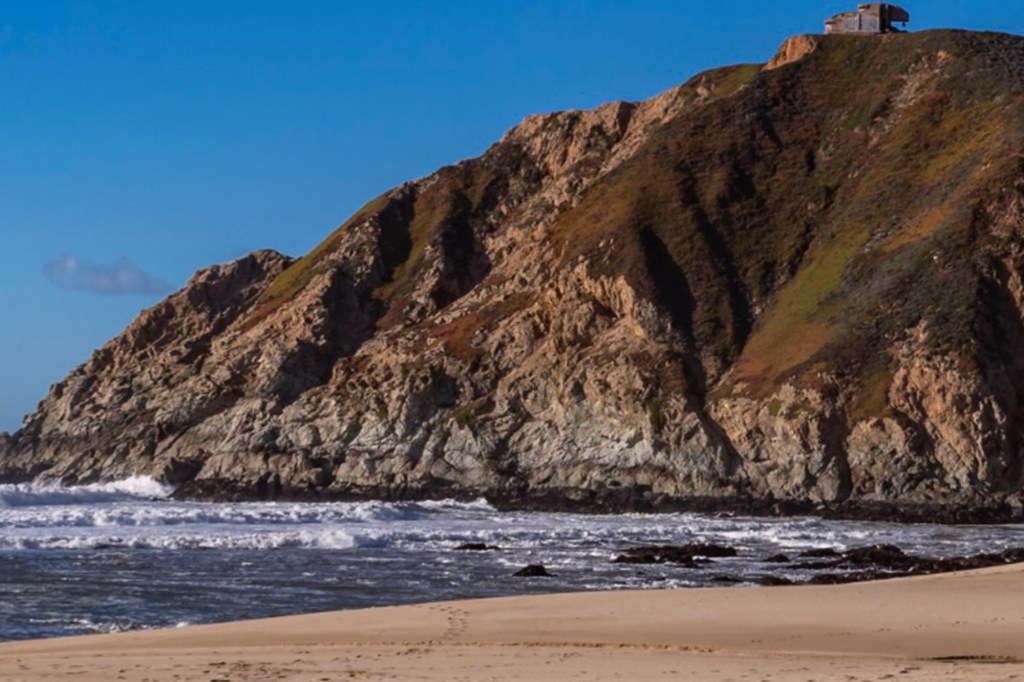 Gray Whale Cove State Beach in San Mateo County, California, showing waves crashing against the rocky shore below a high cliff with a building on top.