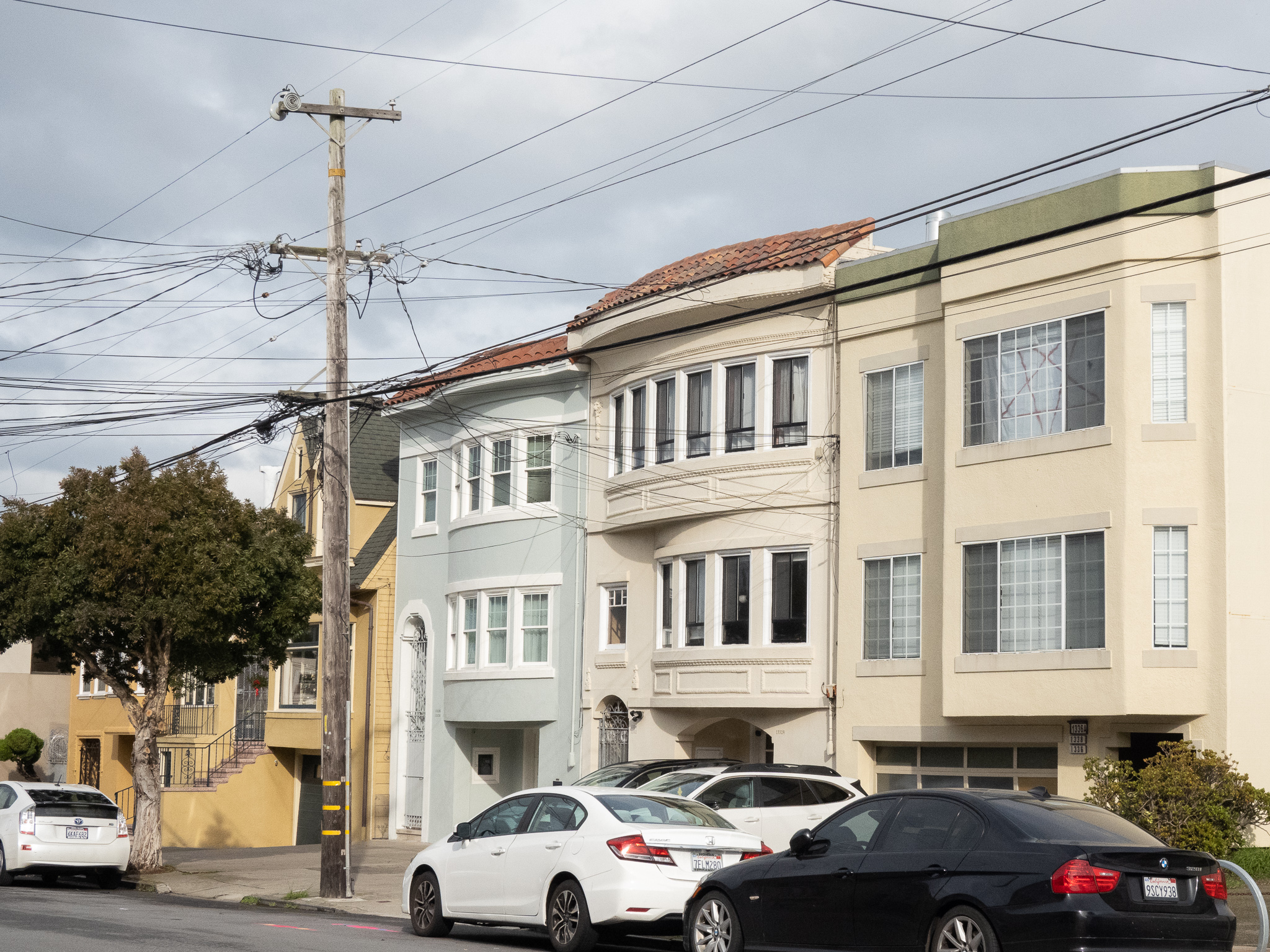 Three cars parked on a residential street in front of multi-story houses, with a utility pole and overhead wires visible.