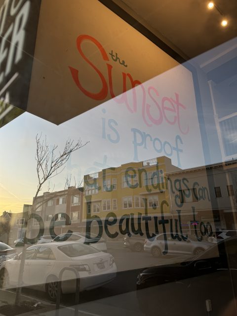 Text painted on a window reads, "The sunset is proof that endings can be beautiful too," with the street and parked cars reflected in the glass.