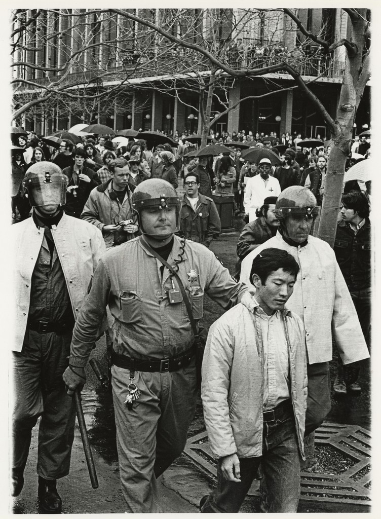 a black-and-white photograph of police wearing helmets walking with an Asian American man. One of the police appears to be carrying a rod and grabbing the arrested activist by the collar.