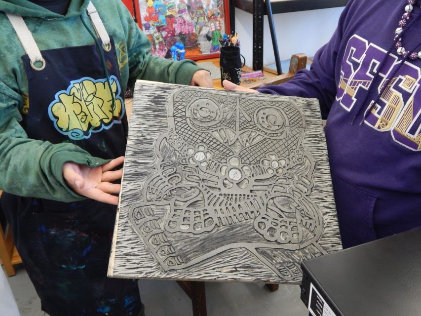 Two people hold a carved printing block featuring an owl design in an art studio. One person gestures towards the block. Art supplies and framed artwork are visible in the background.