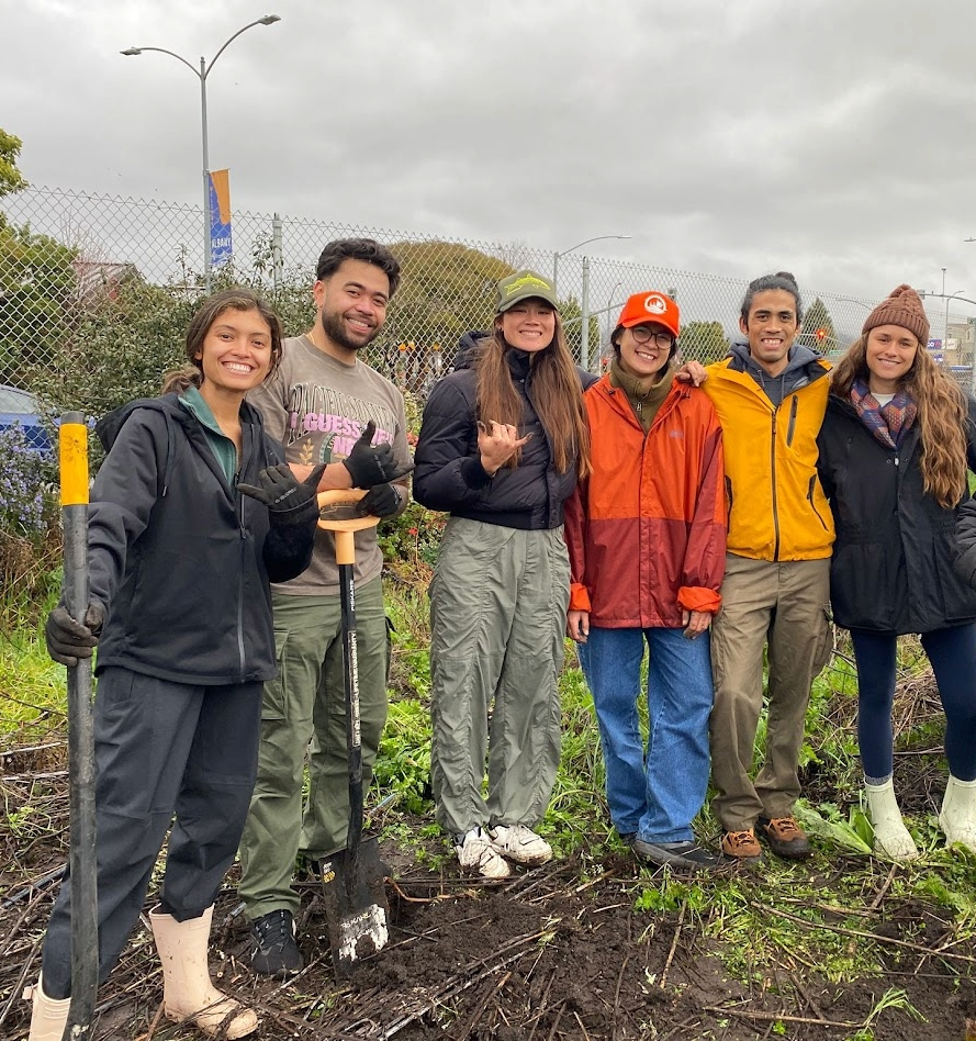 Pacific Islander students in rainy-weather attire stand in a garden with landscaping tools