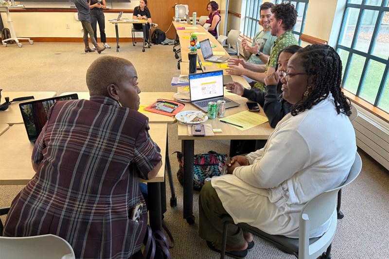 Two participants engage in one-on-one conversation at a table with laptops and materials in a bright classroom setting.