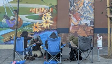 Four people sit in folding chairs on a sidewalk in front of a colorful mural, with scattered belongings nearby, as a marshall stands watchfully at the edge of the scene.