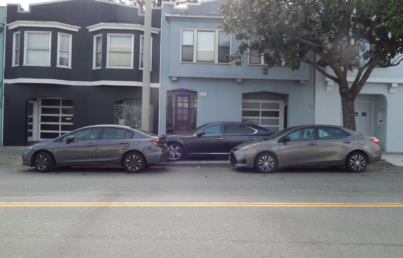 Three cars are parked in a row on a residential street in front of two attached houses, one painted dark gray and the other light blue.
