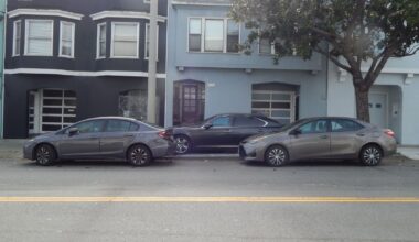 Three cars are parked in a row on a residential street in front of two attached houses, one painted dark gray and the other light blue.