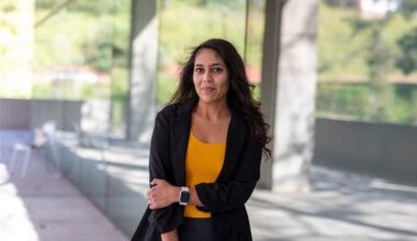 A portrait of a woman standing outdoors by a glass-walled walkway, wearing a black blazer and yellow top, looking at the camera with a neutral expression.