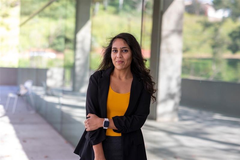 A portrait of a woman standing outdoors by a glass-walled walkway, wearing a black blazer and yellow top, looking at the camera with a neutral expression.