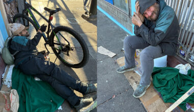 A man sits and lies on the sidewalk near a bicycle and a blanket, surrounded by personal belongings and makeshift bedding on a city street.
