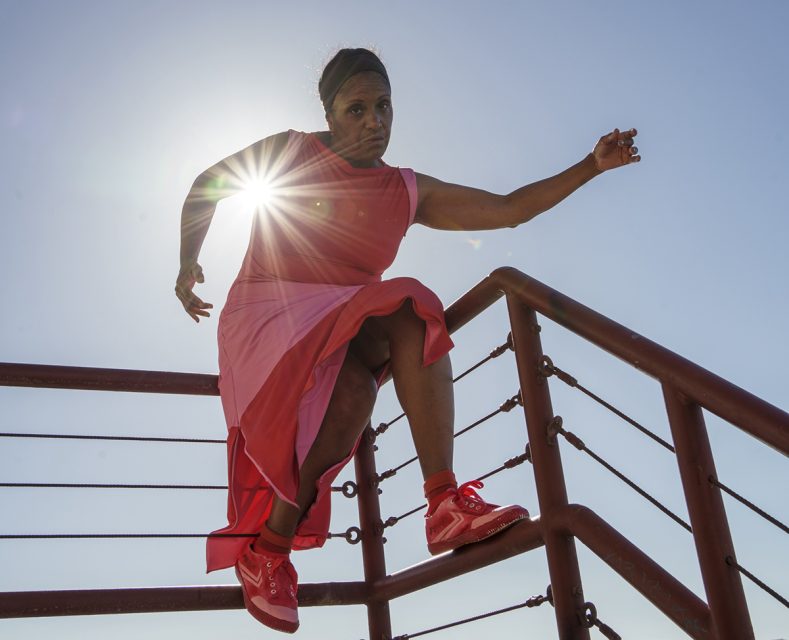 A person wearing a pink dress and red sneakers jumps over a red metal railing with the sun shining behind them.