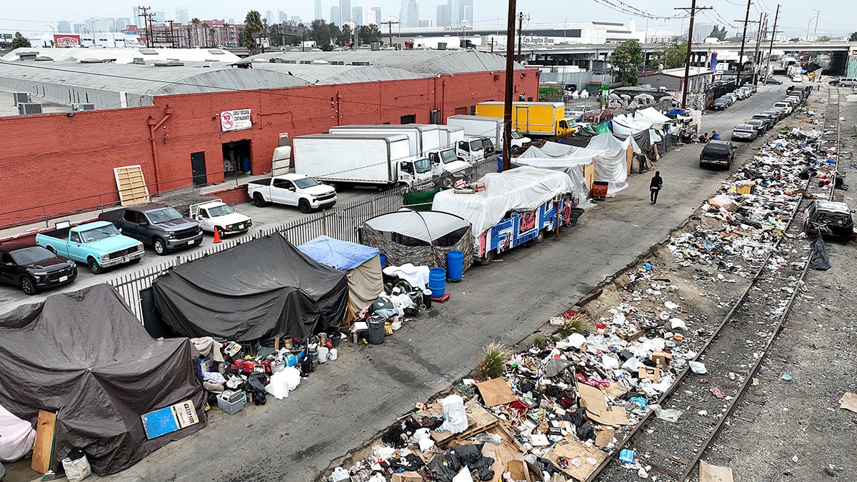 A person walks past large piles of trash inside a sprawling homeless encampment.