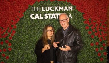 Two guests pose in front of a red rose and greenery photo wall with “The Luckman” and “Cal State LA” signage.