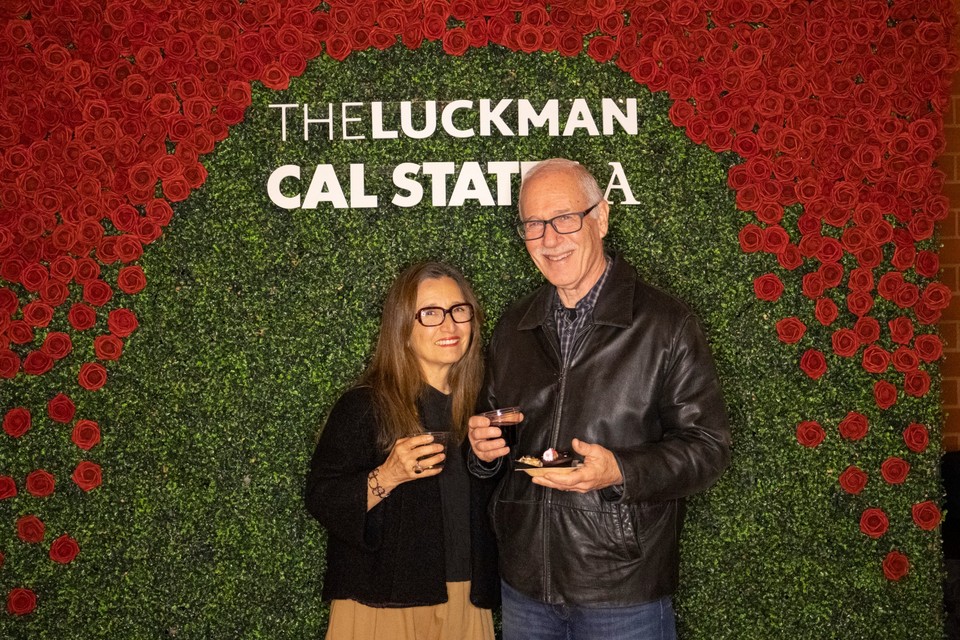 Two guests pose in front of a red rose and greenery photo wall with “The Luckman” and “Cal State LA” signage.