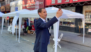 Man in a suit taking a selfie on a city street with market tents and signs in the background.