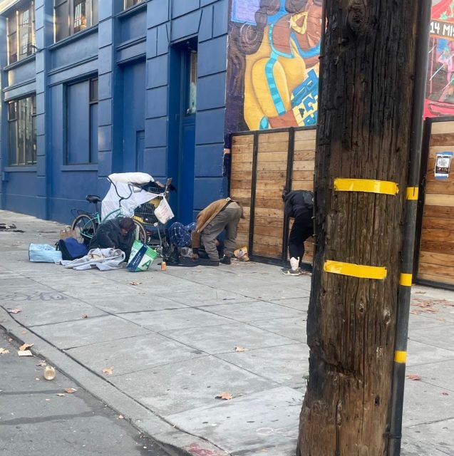 Three people, possibly assisted by a marshall, sort through belongings and bags on a city sidewalk near a blue building with a mural and a wooden fence; a bicycle and scattered items are visible.
