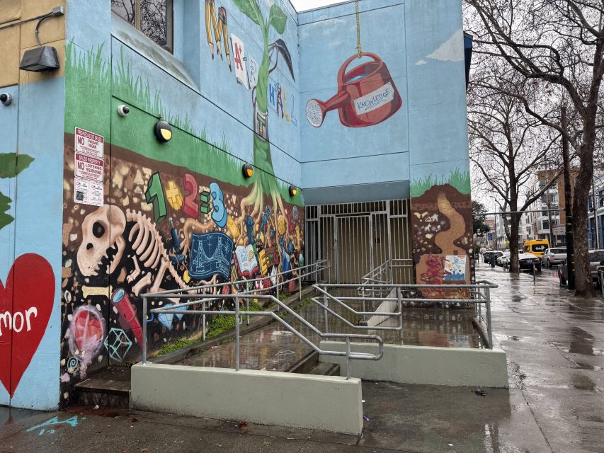 A colorful mural featuring a dinosaur skeleton, numbers, and a watering can decorates the exterior of a building with ramp and handrails in Marshall on a rainy day.