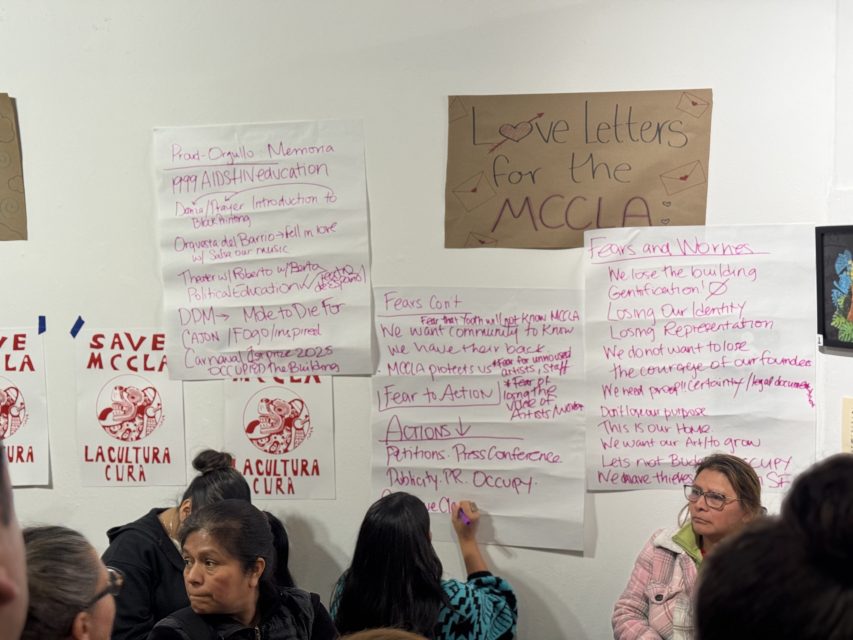 People stand in front of handwritten posters and signs on a wall at a gathering; posters discuss concerns about losing a community center and plans for action.
