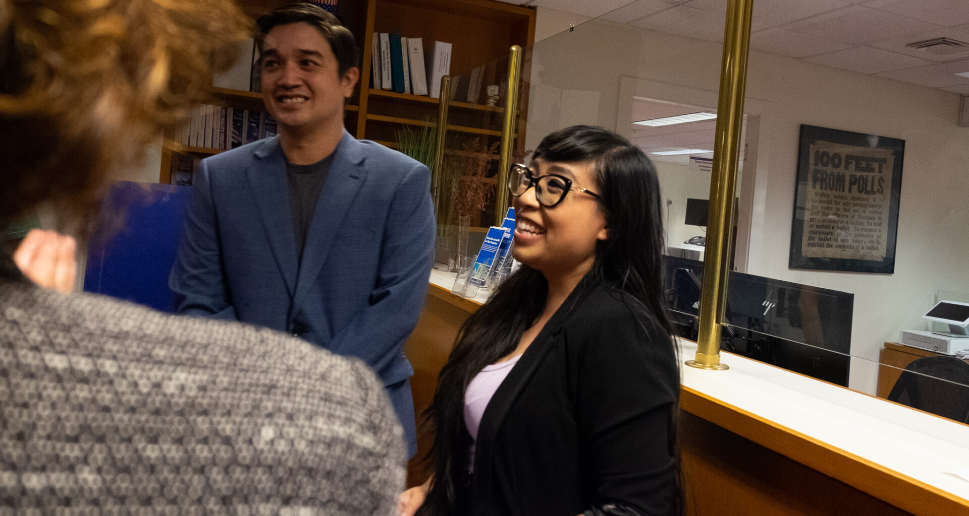 Two people standing and smiling in an office setting, talking to a person whose back is to the camera. A sign in the background reads "100 FEET FROM POLLS.