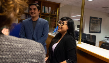 Two people standing and smiling in an office setting, talking to a person whose back is to the camera. A sign in the background reads "100 FEET FROM POLLS.