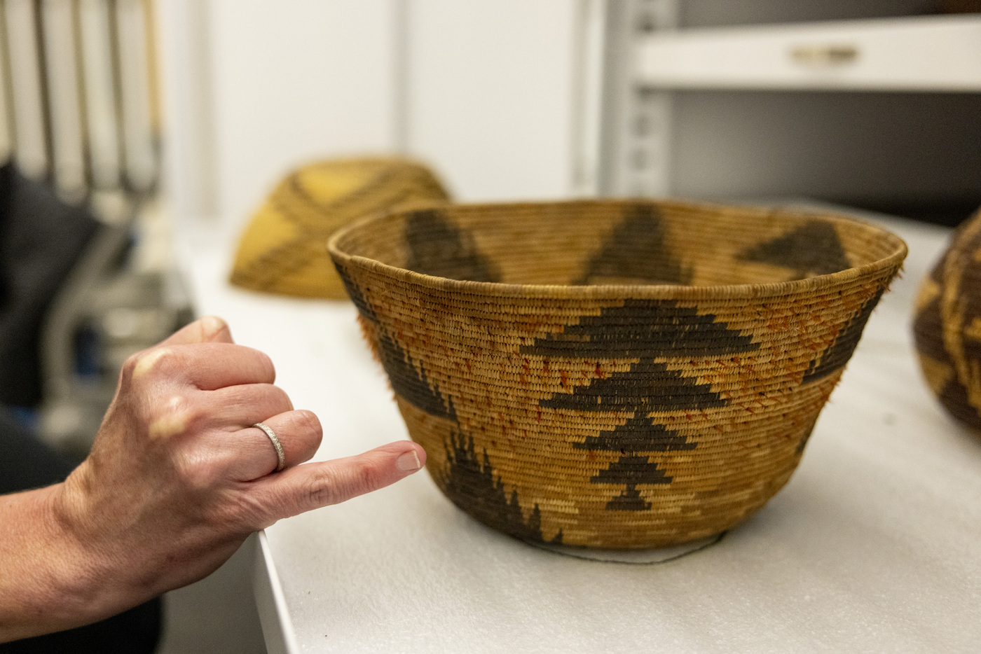 A hand points to the detail of a light-brown woven bowl decorated with dark triangular patterns.