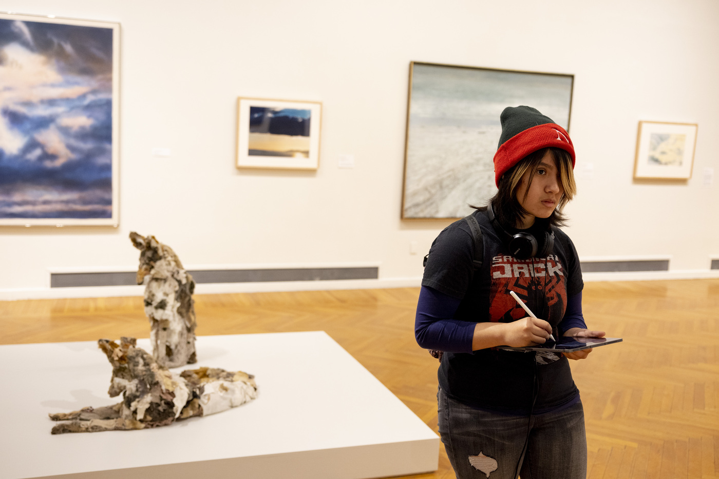 A young person in a black-and-red beanie stands in a gallery filled with artwork, drawing on a tablet. A sculpture on a white podium is to their right.
