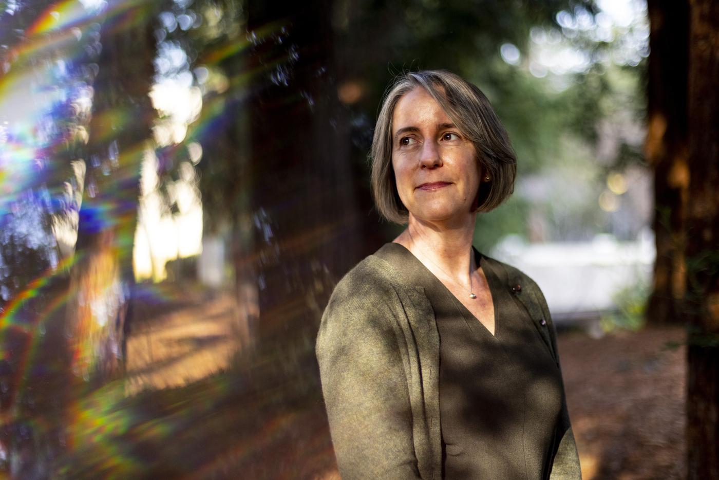 A woman with grey bob haircut, wearing army green t-shirt and a cardigan poses for a half-body portrait in a wooded area. The woman looks off camera and a pleasant facial expression.