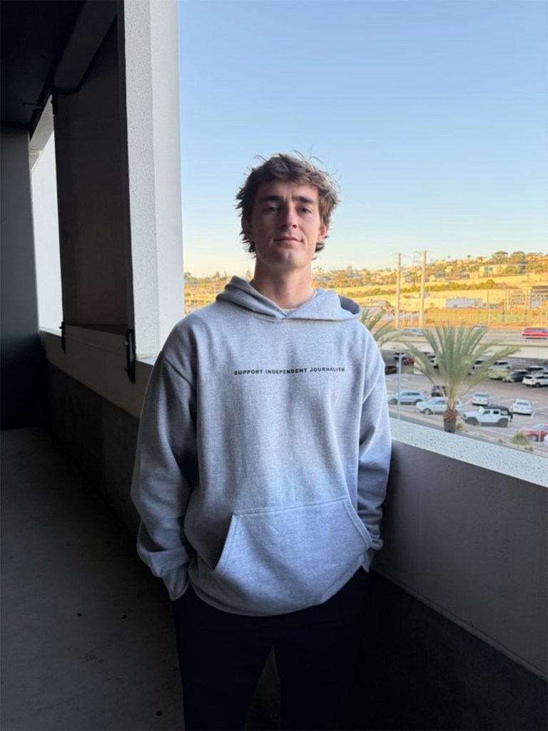 A young man in a grey hoodie with "SUPPORT INDEPENDENT JOURNALISM" printed on it stands on a balcony overlooking a city.