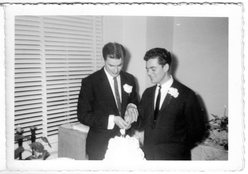 a black-and-white photograph of two men in tuxedos and boutonnieres cutting a wedding cake