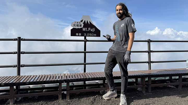 A young man in athletic wear stands by the railing and bench of a lookout point with his right thumb pointing toward a sign that reads Mount Fuji, 3250m