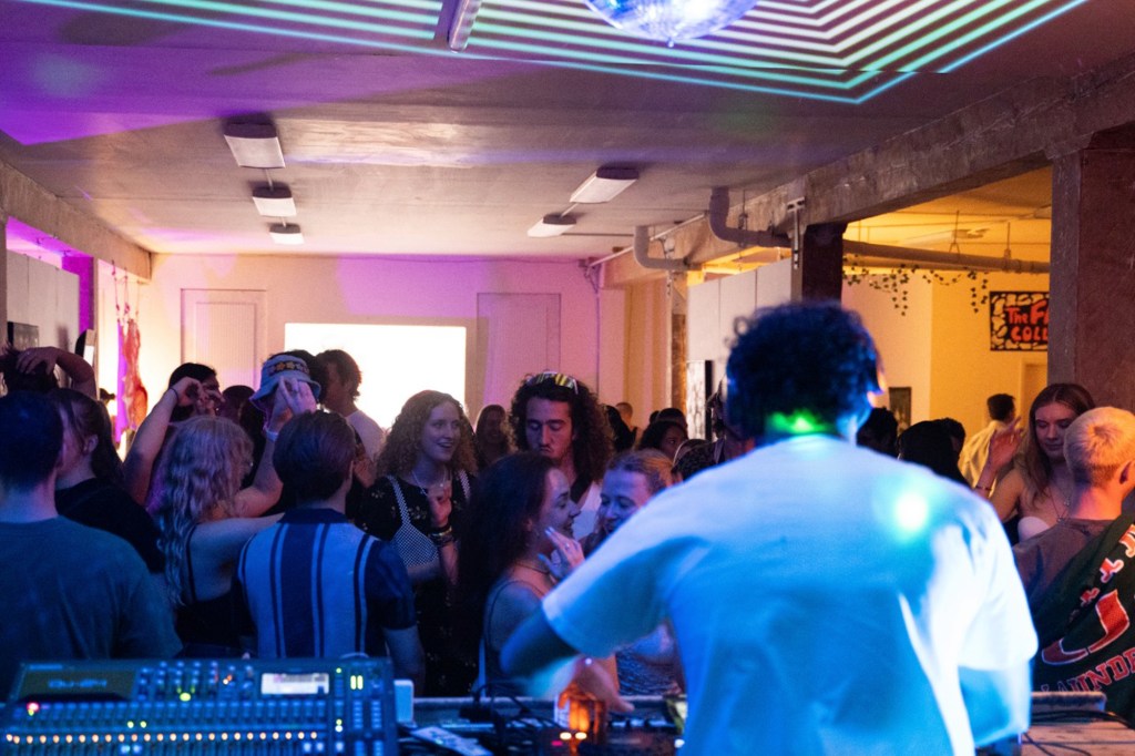 A DJ playing at a party with a crowd of people dancing and a disco ball reflecting light patterns on the ceiling.